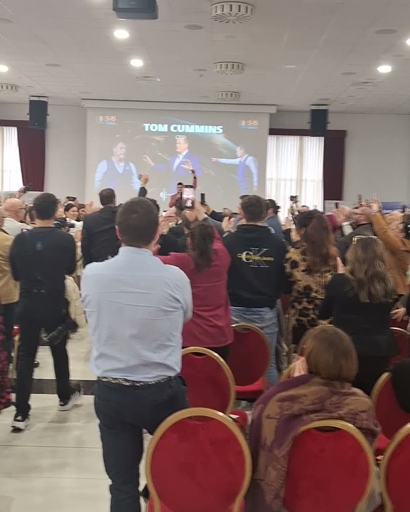 A standing audience raising phones toward a stage screen reading Tom Cummins